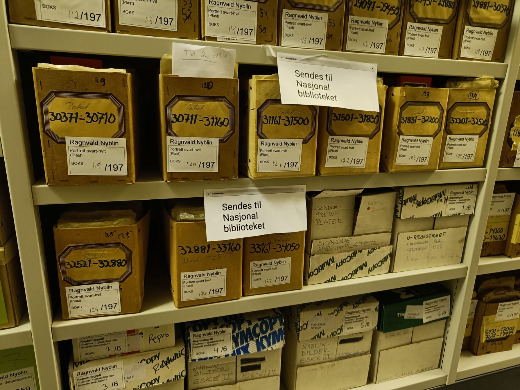 Bilde av gamle arkivkasser i hyller. På en lapp står det: Sender til nasjonalbiblioteket