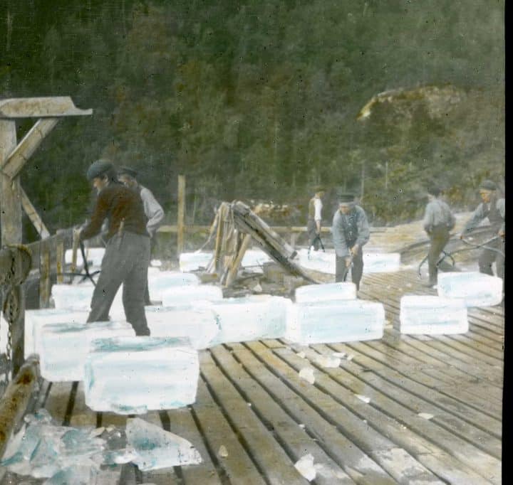 Et gammelt bilde med svake farger viser fire menn som hogger ut isblokker. Blokkene ligger på en treplatting