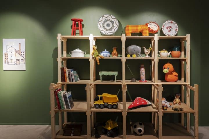 Utstillingen Koppen på Porselensmuseet. Bildet viserr en hylle med leker, bøker og porselen. Furuhylle mot en grønn vegg. Lekene er fargerike.