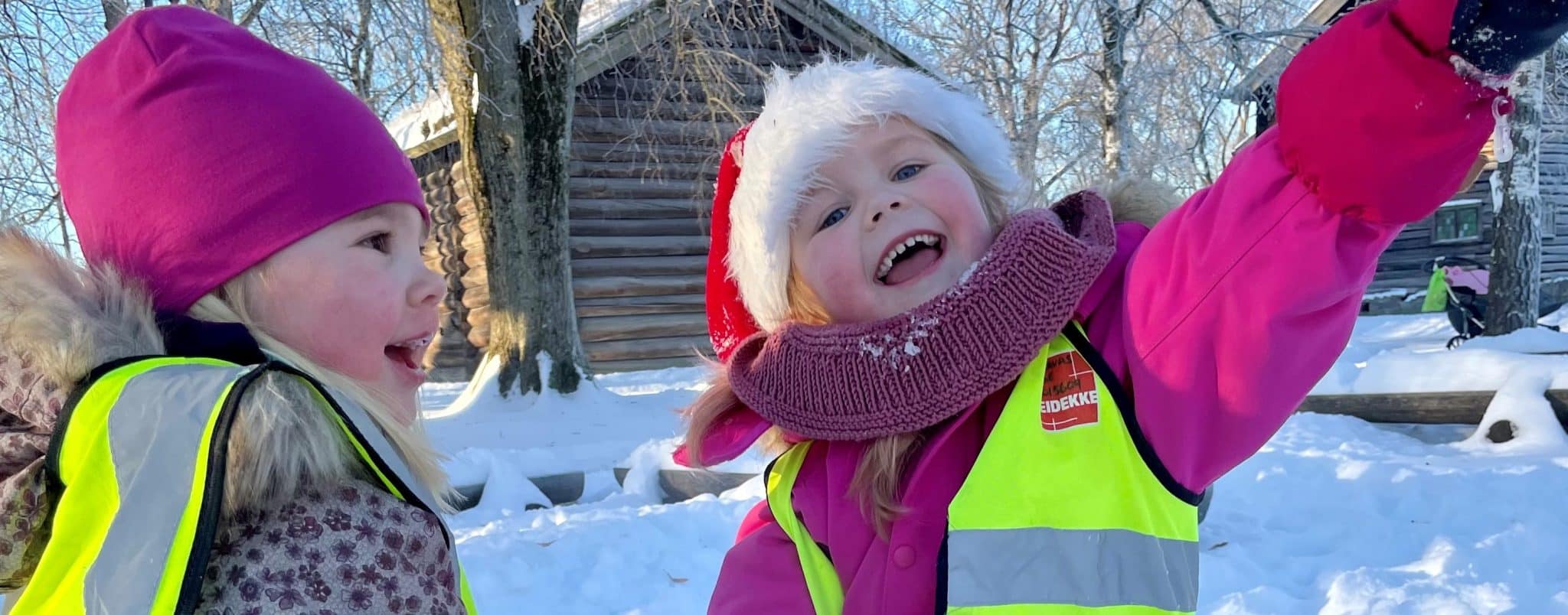 To glade barn på lekeplassen i Brekkeparken. Tømmerstuer og snø vises i bakgrunnen