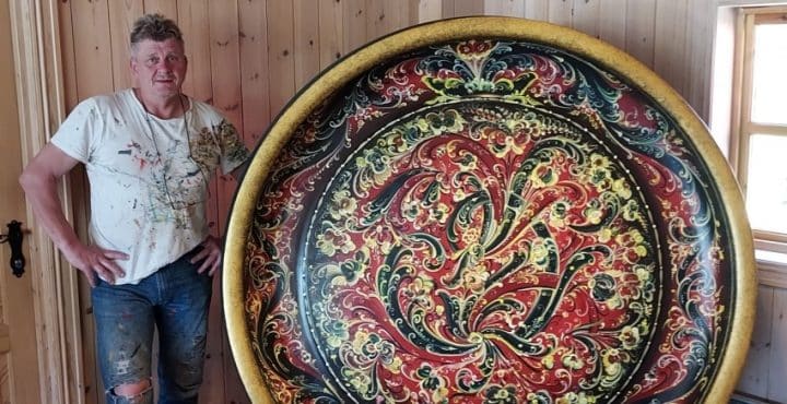 Kurs i rosemaling. Maleren selv poserer foran en enorm, rosemalt bolle. Bollen er like høy som mannen ved siden av.