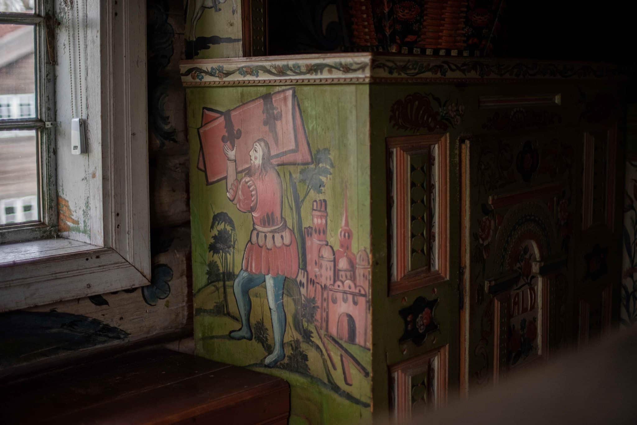 Kroneseng i andre etasje på Rambergstua. Detaljbilde av enden på senga. Senga er malt i grønn bunnfarge med et bibelsk motiv. En mann bærer en stor bok og i bakgrunnen ser vi en moskelignende bygning fra østen