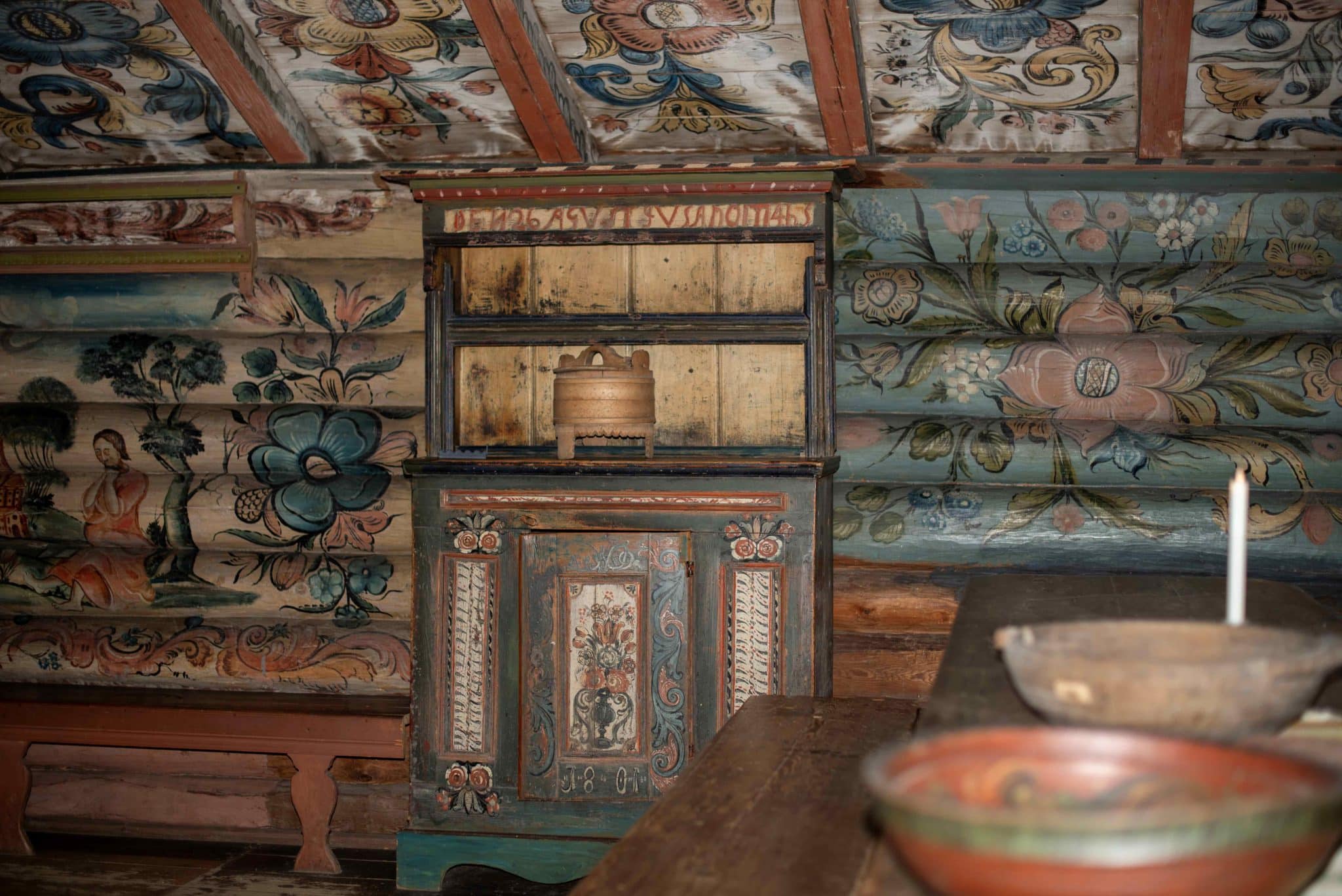 Rambergstua innvendig, gjestestua i andre etasje. Et rosemalt framskap i midten som står plassert til en rosemalt tømmervegg. I forkant skimter vi to rosemalte ølboller på et langbord