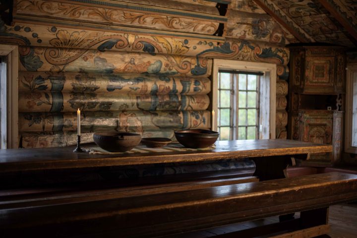 Rambergstua innvendig med langbord. Rikt dekorerte tømmervegger, rosemalt av Ola Hansson. Foran veggen står et langbord med to store og en liten ølbolle, og to lystestaker på bordet.