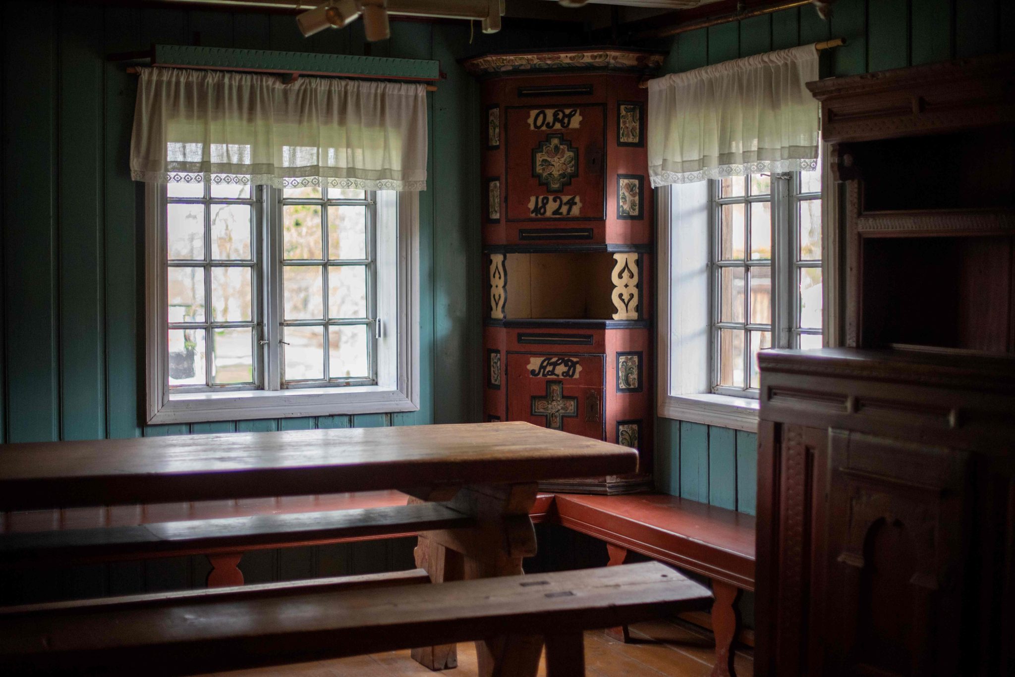 Rambergstua innvendig med langbord og to smårutete vinduer. Rosemalt kroskap i hjørnet mellom de to vinduene