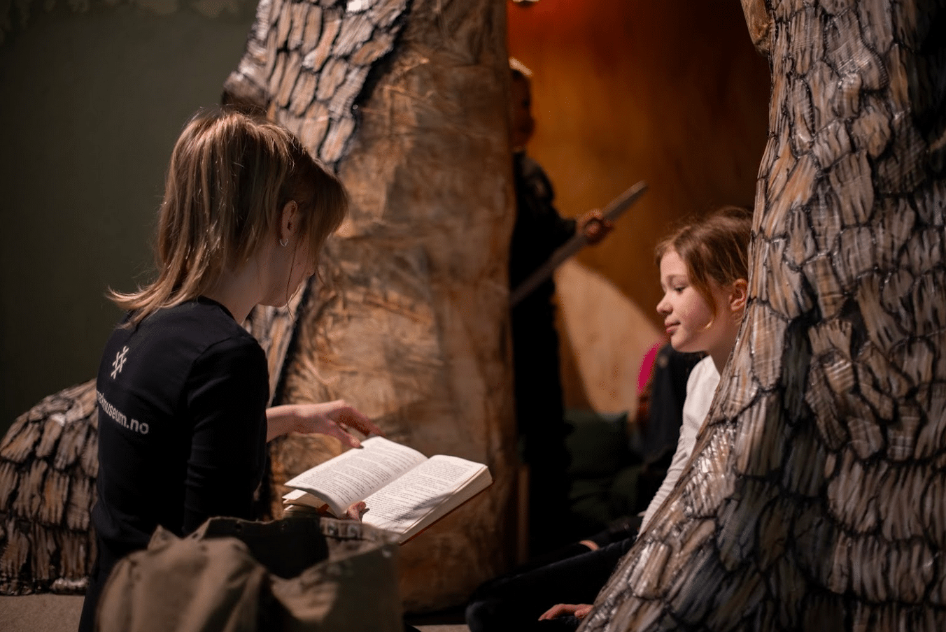 Eventyrstund i Theodor Kittelsens verden. En omviser fra Telemark museum sitter og leser i en bok for barn som hører på. Rundt dem er kulisser i utstillingen Eventyrlige Kittelsen.