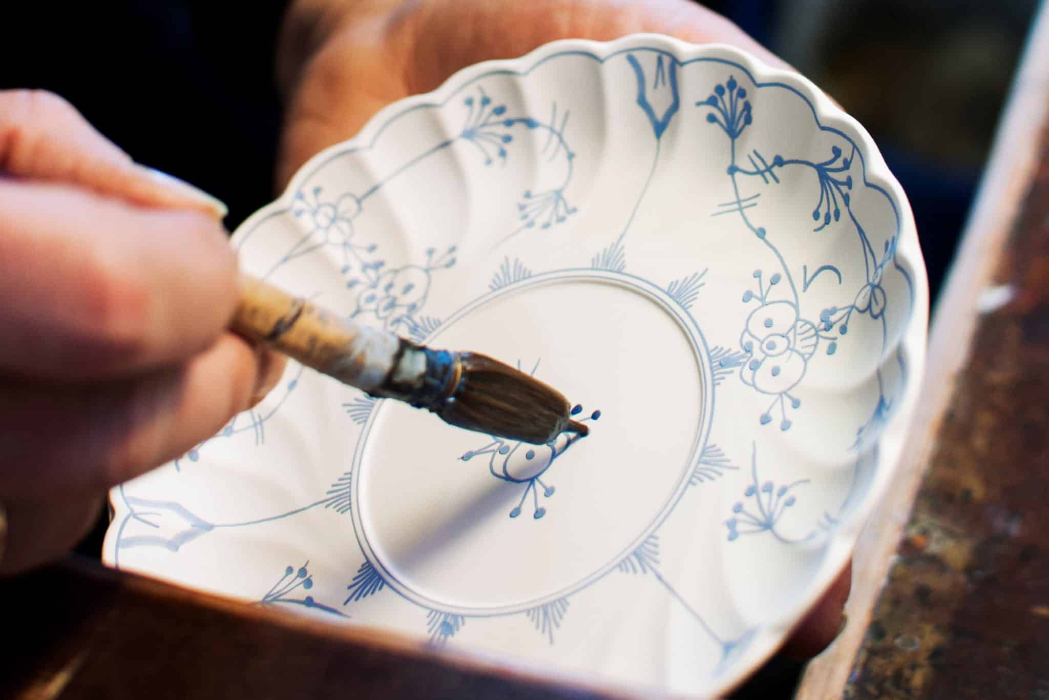 Maling av stråmønsteret på Porsgrund Porselænsfabrik. En hånd holder en pensel og maler blått strå mønster på en hvit tallerken