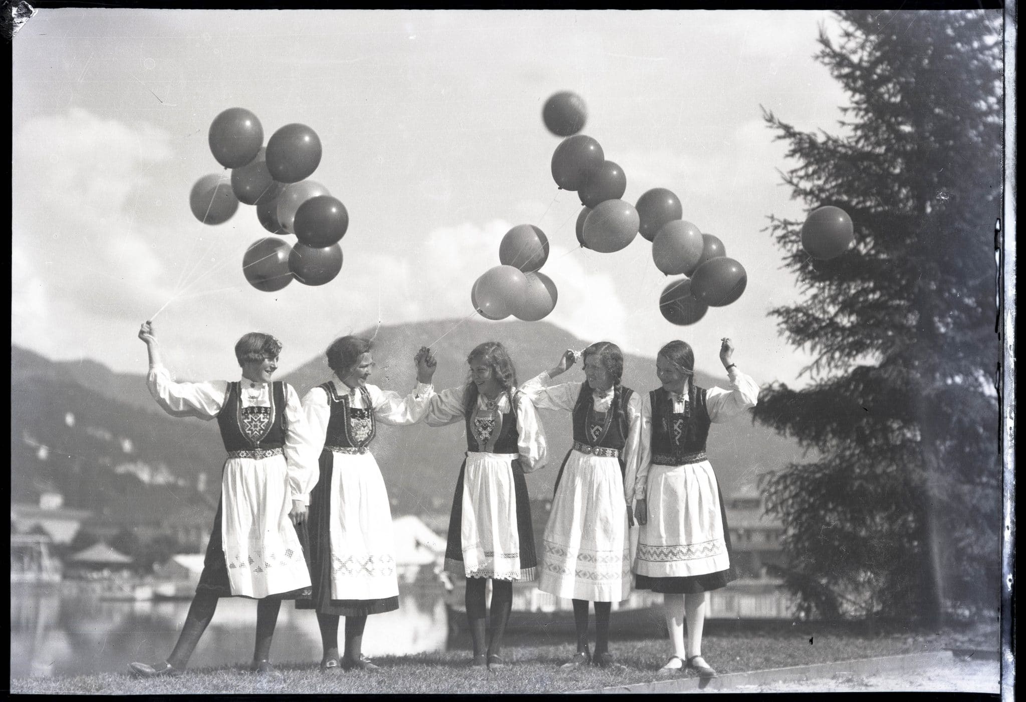 Fem unge kvinner i folkedrakt holder på ballonger og ler. Fotograf: Jensen Atelier, sort-hvitt bilde