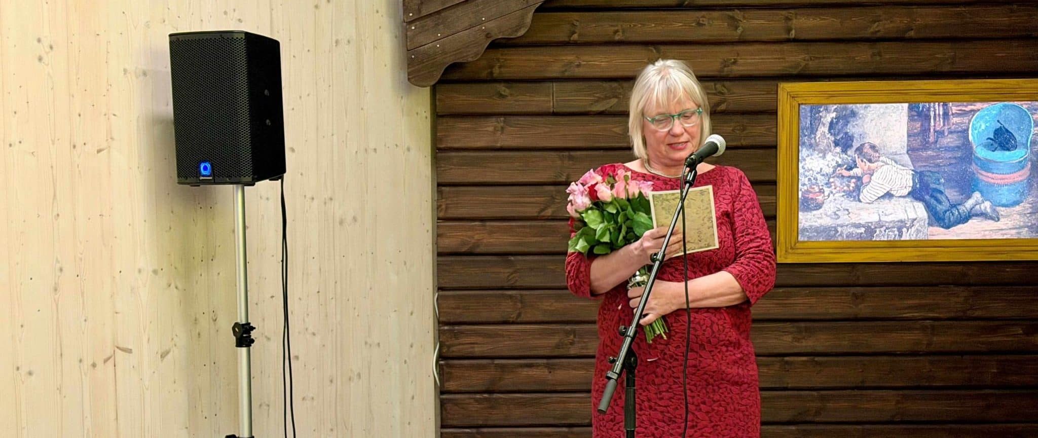 Unni Værstad står og leser dikt. I bakgrunnen ser vi en del av en tømmerhytte.