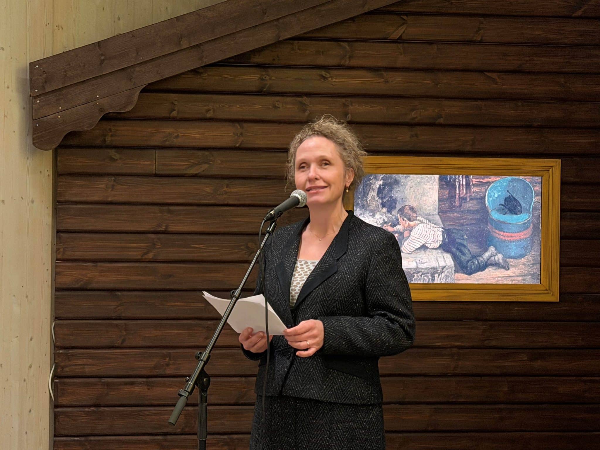 Christine Hermansen snakker i mikrofonen, stående foran en panlet, brun hyttevegg.