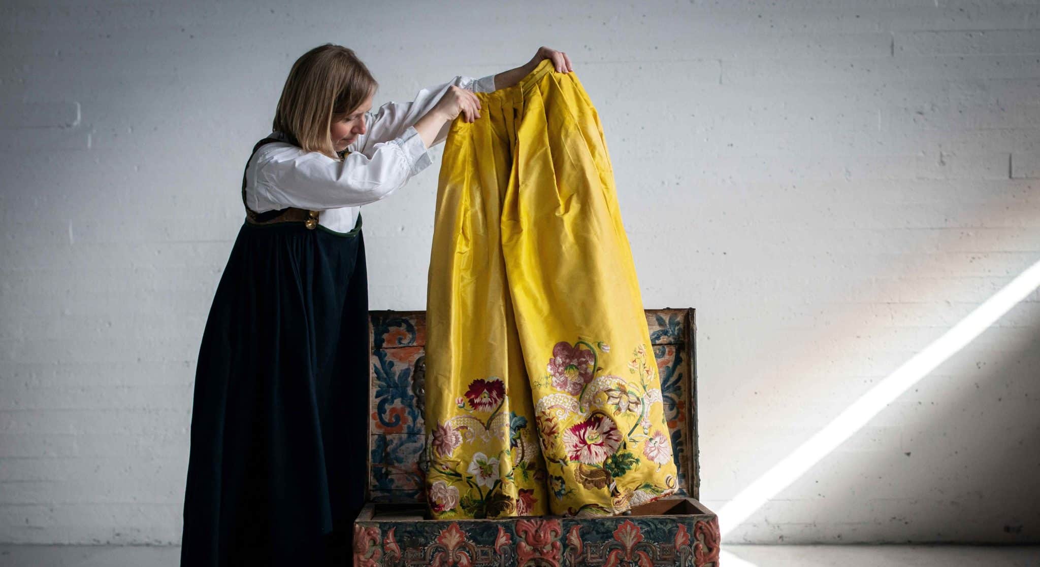 Den gule kjolen i utstillingen Alt dette er ditt. En dame i stakk og hvit skjorte løfter opp et gult skjørt med flott mønster nederst opp fra en rosemalt kiste