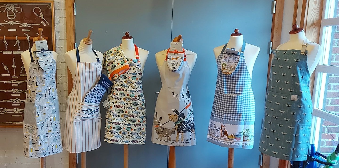 Fra museumsbutikken på Berg-Kragerø Museum. Fem ulike forklær med mønster står på rekke på byster. Det er fiskemønster, hundemønster og andre mønster på forklærne.