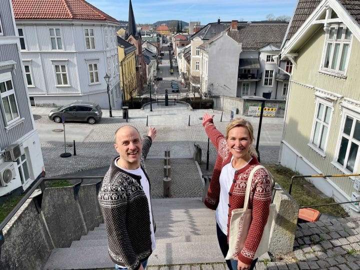 Ibsen walking tour in Skien. A man and a woman pose on either side of the Henrik Ibsen bust at Sorrento Square in Skien. They raise their arms, pointing down the street that leads all the way to the yellow wooden house on the hilltop across the town.