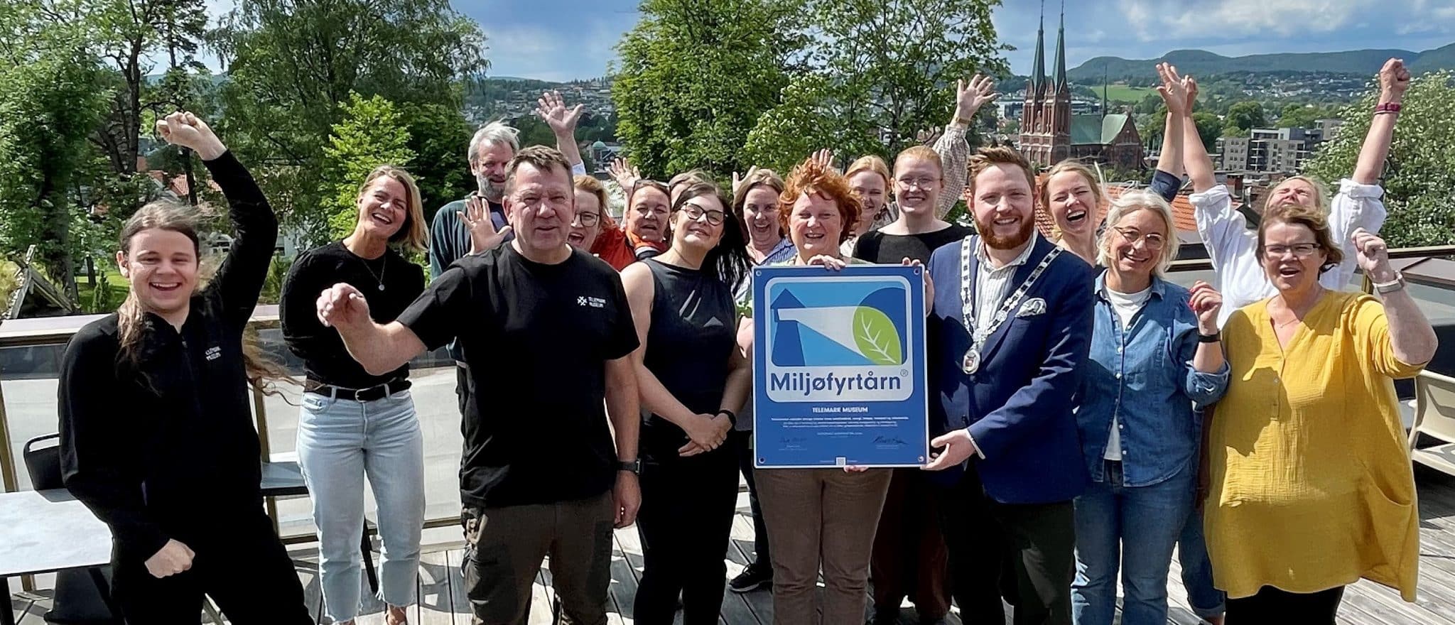 Telemark museum sine ansatte jubler på takterrassen. Bedriften er blitt miljøsertifisert og skiltet vises i forgrunnen. Kirkespirene på SKien kirke vises i bakgrunnen.
