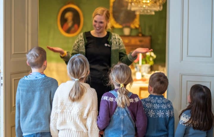 Turist i egen by, omvisning i hovedhuset i Brekkeparken. En kvinnelig guide i grønn Telemark museumskofte står foran fem barn og forteller om hovedhuset. I bakgrunnen ser vi en herskapelig sal med grønne vegger og portretter på veggene.